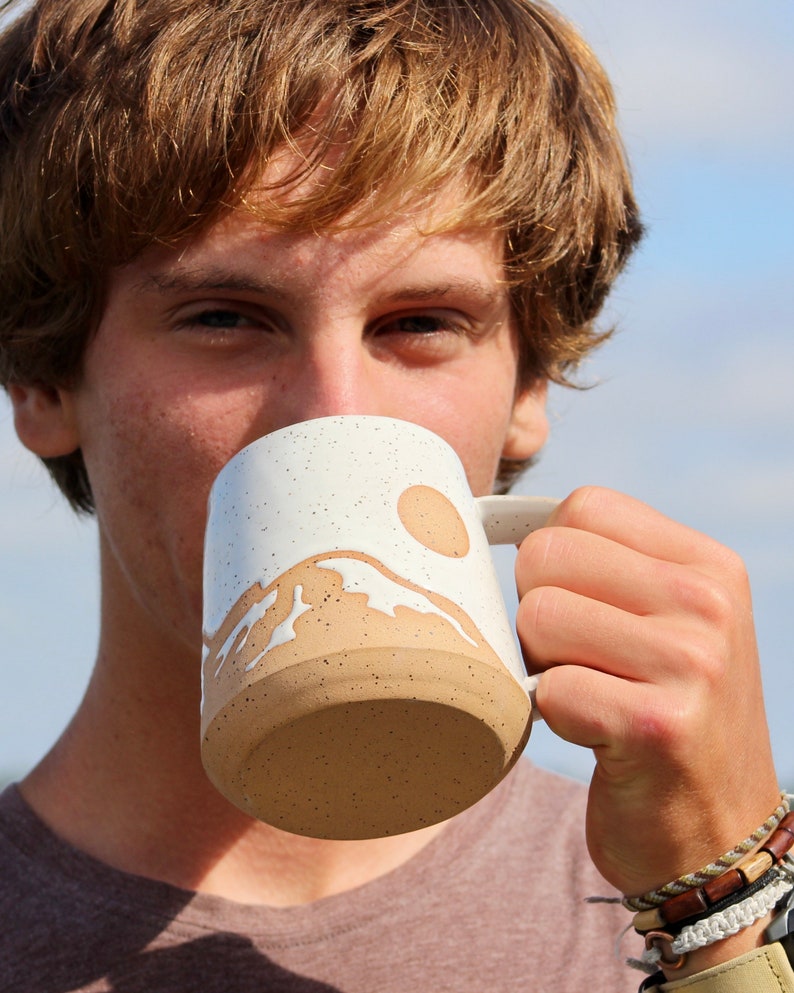 May include: A white ceramic mug with a brown speckled glaze. The mug features a white mountain range design with a brown sun and waves.