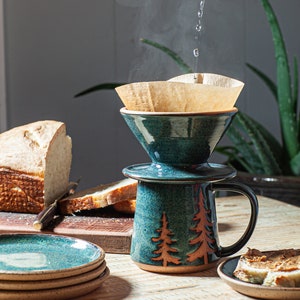 May include: A teal blue ceramic coffee pot with a brown paper filter and a wooden handle. The pot is decorated with a brown tree design. A white loaf of bread and a knife are in the background.