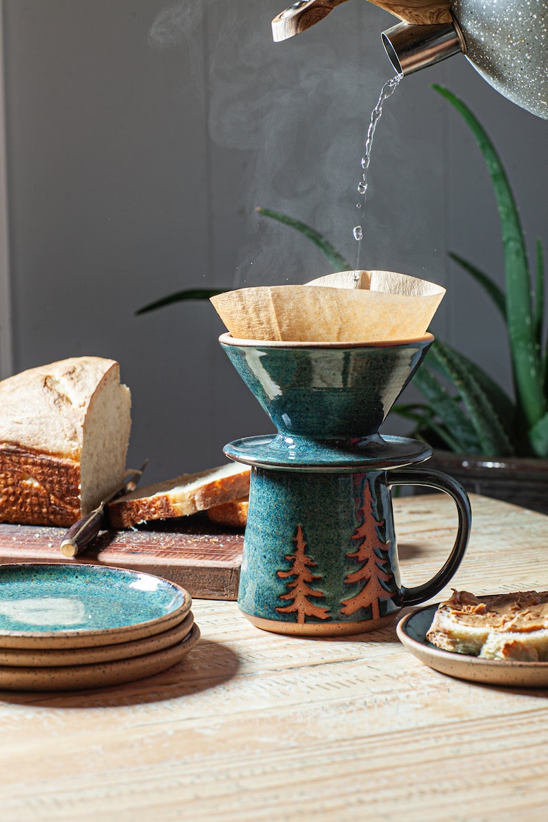 May include: A teal blue ceramic coffee pot with a brown paper filter and a wooden handle. The pot is decorated with a brown tree design. A white loaf of bread and a knife are in the background.