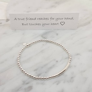 May include: A silver beaded bracelet with a small heart charm and a silver bar accent. The bracelet is on a white marble surface. The text on the card reads: "A true friend reaches for your hand, But touches your heart".