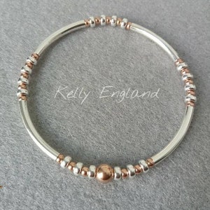 May include: A silver and rose gold bracelet. The bracelet has a silver tube design with rose gold and silver beads. A single rose gold bead is in the centre. The text "Kelly England" is printed on the background.