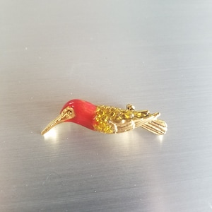 May include: A gold-toned hummingbird brooch with a red head and a gold-colored body. The bird's beak is long and slender. The brooch is a decorative accessory.