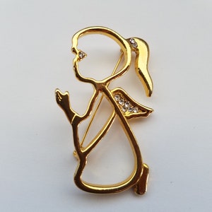 May include: A gold-colored metal brooch in the shape of a kneeling angel. The angel has a ponytail and small crystal accents on the wings and face. The brooch is a detailed, decorative accessory.