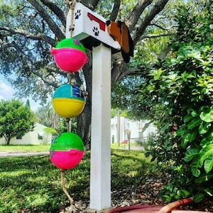 May include: A white mailbox with a dog head design, painted in brown, red, and black. The mailbox is attached to a white post with three colorful, round, floating buoys hanging from a rope.
