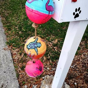 May include: Three painted, round, hanging ornaments with a rope attached. The top ornament is pink and blue with a flamingo painted on it. The middle ornament is yellow and orange with a turtle painted on it. The bottom ornament is pink with a floral design.