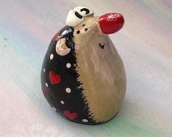 May include: A whimsical, handmade ceramic figurine of a creature with a red nose and a black and tan body. The black side is decorated with red hearts and white polka dots. It has a small white hat.