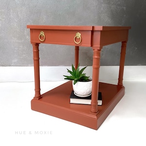 May include: A terracotta-colored side table with a drawer and brass ring pulls. The table has four decorative legs and a lower shelf. A white patterned pot with a green plant sits on the shelf, along with a book. The text "HUE & MOXIE" is at the bottom.