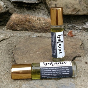 May include: Two gold roller bottles with black labels that read "balance". The labels list the ingredients: Organic Grapeseed Oil, Therapeutic Grade Tangerine, Orange, Lavender, Clove, Ylang Ylang, Patchouli, & Blue Tansy Essential Oils 10ml. Directions: Roll on inside of wrists, behind ears, bottom of feet as needed. www.WonderfullyMadeLove