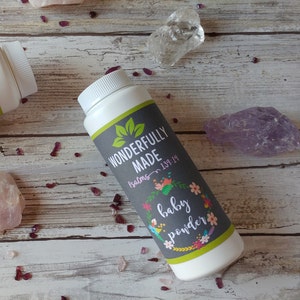 May include: Two white plastic bottles of baby powder with green and gray labels. The labels have text that reads "Wonderfully Made" and "Baby Powder". The bottles are on a wooden surface with pink and purple crystals.