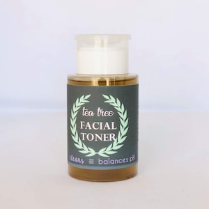May include: A clear glass bottle with a white pump top and a black label. The label says "tea tree facial toner" with a green laurel wreath around the text. The label also says "cleans and balances pH".