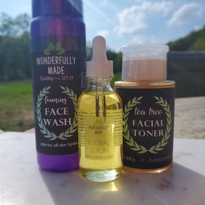 May include: Three skincare products are displayed: a purple foaming face wash, a clear floral serum, and a tea tree facial toner. The face wash and toner are in cylindrical bottles with black labels. The serum is in a small glass bottle with a dropper.
