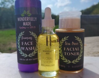 May include: Three skincare products are displayed: a purple foaming face wash, a clear floral serum, and a tea tree facial toner. The face wash and toner are in cylindrical bottles with black labels. The serum is in a small glass bottle with a dropper.