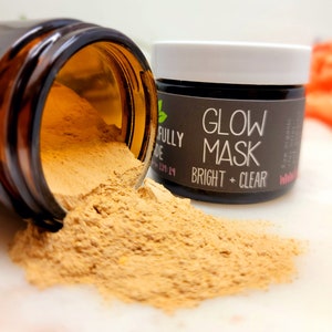 May include: A brown glass jar with a white lid is spilling out a light brown powder. The jar is labeled "Glow Mask Bright + Clear" with a green leaf logo.