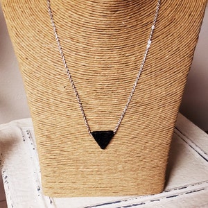 May include: A silver chain necklace with a black triangular pendant. The necklace is displayed on a tan bust form. The bust is sitting on a white, distressed wooden surface.