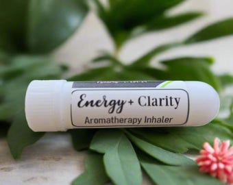 May include: A white aromatherapy inhaler with the words "Energy + Clarity" printed on a label. The inhaler is surrounded by green leaves and a small pink flower. The product is designed for aromatherapy.
