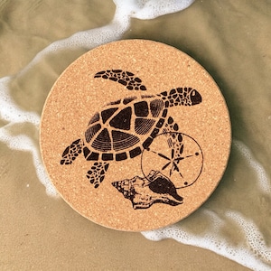 May include: A round cork trivet with a black silhouette of a sea turtle, a seashell, and a sand dollar. The trivet is on a sandy background with white foam.