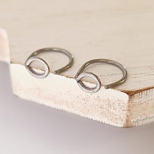 May include: Two silver-colored hoop earrings with a teardrop-shaped design. The earrings are placed on a light-colored wooden surface. The earrings are small and delicate, with a simple, minimalist style.