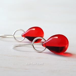 May include: A pair of teardrop-shaped earrings. Each earring features a vibrant red, translucent glass bead with a dark red base, suspended from a silver-colored, circular hook. The earrings are displayed on a light-colored surface.
