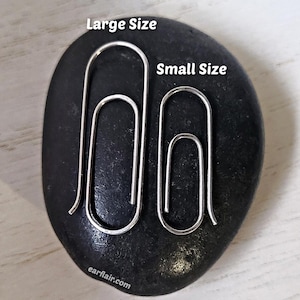 May include: Two silver-colored paperclip-shaped earrings, one large and one small, are displayed on a dark, oval-shaped stone. The text "Large Size" and "Small Size" are above the respective earrings. The website "earflair.com" is visible at the bottom.