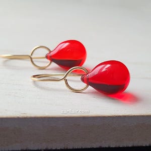 May include: A pair of gold-colored earrings featuring teardrop-shaped red glass pendants. The earrings have a simple, elegant design with a small loop connecting the pendant to the ear wire. The red glass has a dark red hue at the bottom.