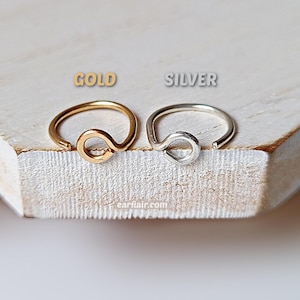 May include: Two nose rings, one gold and one silver, are displayed on a light-colored wooden surface. The gold ring is on the left, and the silver ring is on the right. Each ring has a looped design.