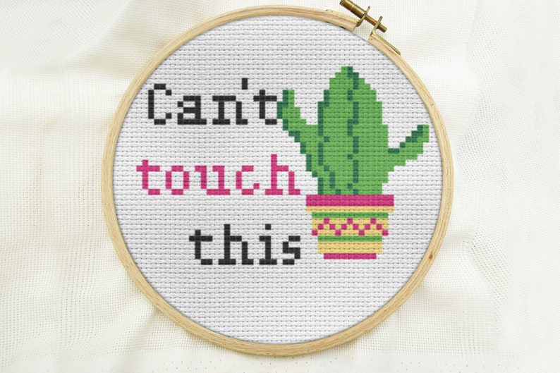 K&ouml;nnte beinhalten: Ein wei&szlig;er Stickrahmen mit einem gr&uuml;nen Kaktus in einem gelben und rosafarbenen Topf. Der Text "Can't touch this" ist in Schwarz und Rosa gestickt.