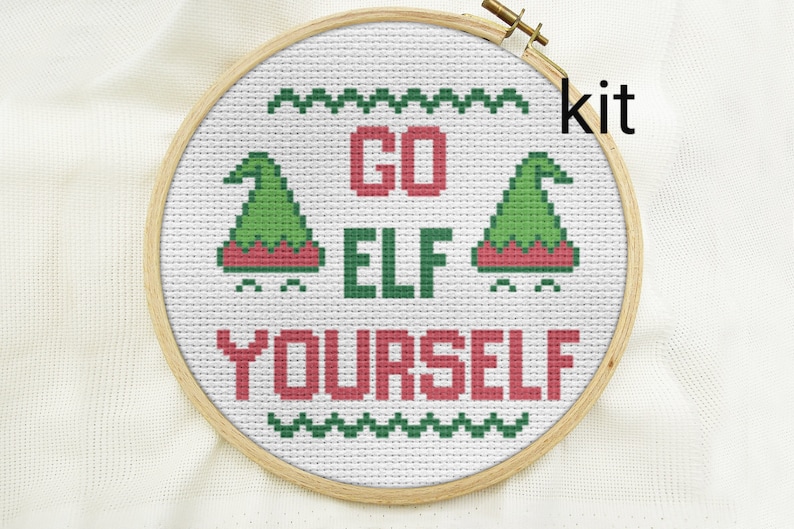Puede incluir: Un kit de punto de cruz con un fondo blanco y bordado verde y rojo. El dise&ntilde;o dice "Go Elf Yourself" con dos gorros de elfo a cada lado del texto.