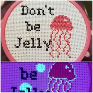 May include: A white embroidery hoop with a pink rim, featuring a cross-stitch design of a pink jellyfish with the text "Don't be jelly". The jellyfish glows under a black light, with the text and jellyfish glowing in a light blue color.