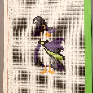 May include: A needlepoint canvas featuring a cross-stitched penguin dressed as a witch. The penguin wears a black and purple pointed hat with a yellow buckle, a purple cape with a lime green sash, and orange feet. The canvas has a bright green border.