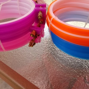 Puede incluir: Dos aros de bordado de pl&aacute;stico de colores. Uno es morado y rosa, y el otro es azul y naranja. Cada aro tiene un mecanismo de ajuste met&aacute;lico y est&aacute; asegurado con una brida blanca. Los aros est&aacute;n sobre una superficie de vidrio.