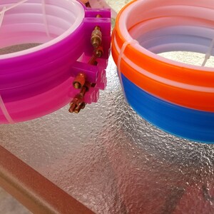 K&ouml;nnte beinhalten: Zwei S&auml;tze Stickrahmen, ein Satz ist lila, rosa und wei&szlig;, der andere Satz ist orange, blau und wei&szlig;.