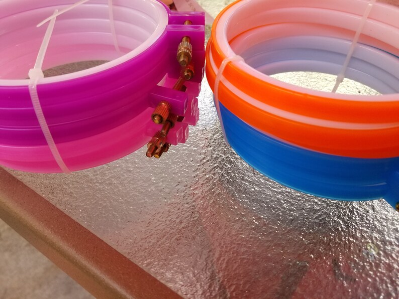 K&ouml;nnte beinhalten: Zwei S&auml;tze Stickrahmen, ein Satz ist lila, rosa und wei&szlig;, der andere Satz ist orange, blau und wei&szlig;.