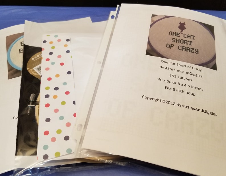 K&ouml;nnte beinhalten: Ein wei&szlig;es Blatt Papier mit dem Text "One Cat Short of Crazy" in schwarzer Schrift. Der Text ist von einem lila und wei&szlig;en Stickrahmen umgeben. Das Papier enth&auml;lt auch den Text "By 4StitchesAndGiggles", "395 stitches", "40 x 60 or 3 x 4.5 inches", "Fits 6 inch hoop", und "Copyright&copy;2018 4StitchesAndGiggles".