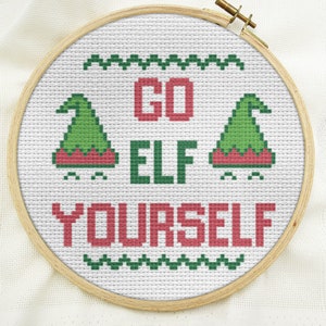 Puede incluir: Un aro de bordado blanco con un dise&ntilde;o de punto de cruz que dice "GO ELF YOURSELF". El dise&ntilde;o presenta dos gorros de elfo verdes con ribete rojo y un borde verde y blanco.