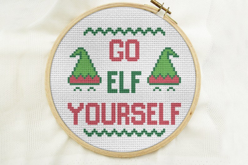 Puede incluir: Un aro de bordado blanco con un dise&ntilde;o de punto de cruz que dice "GO ELF YOURSELF". El dise&ntilde;o presenta dos gorros de elfo verdes con ribete rojo y un borde verde y blanco.