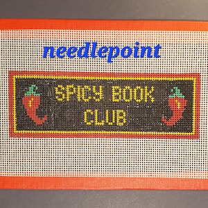 May include: Needlepoint canvas with the text "needlepoint" in blue. The center features a black rectangle with "SPICY BOOK CLUB" in yellow, flanked by red chili pepper designs. The canvas has a white grid pattern with an orange border.
