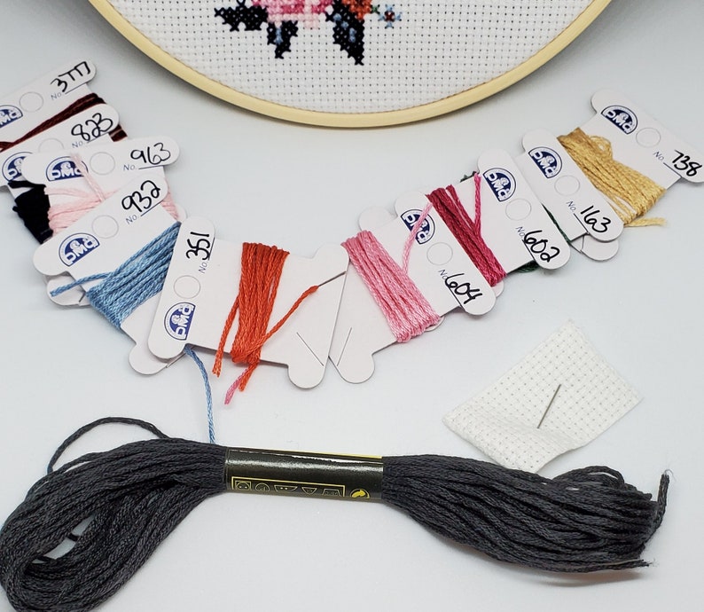 Puede incluir: Surtido de carretes de hilo de bordar DMC en varios colores, incluyendo rojo, rosa, azul, verde y marr&oacute;n. Los carretes est&aacute;n etiquetados con los n&uacute;meros DMC. Tambi&eacute;n se incluye un peque&ntilde;o trozo de tela Aida blanca con una aguja.