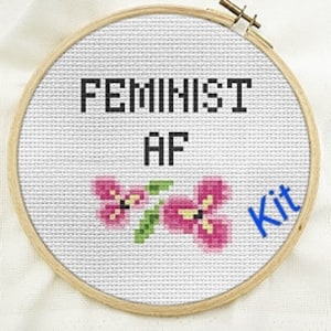 May include: A white embroidery hoop with the text "Feminist AF" and two pink flowers. The text "Kit" is written in blue in the lower right corner.
