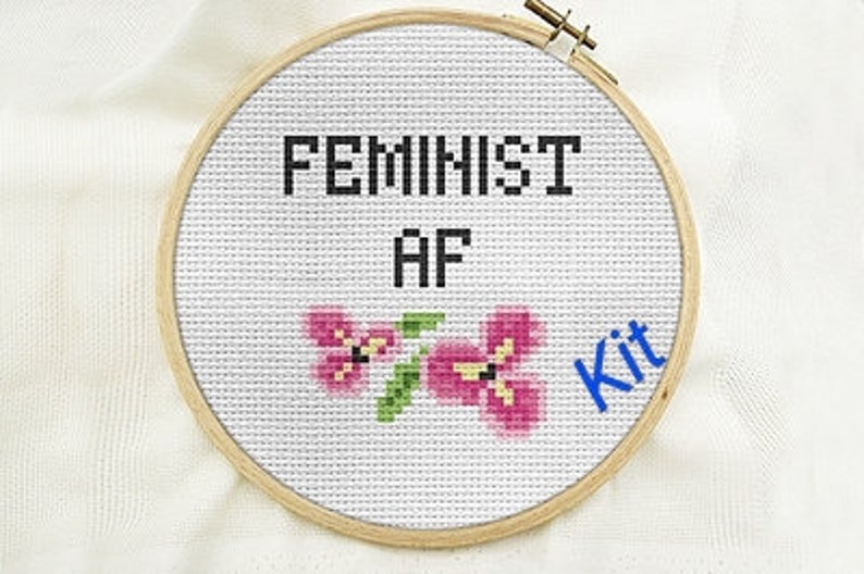 May include: A white embroidery hoop with the text "Feminist AF" and two pink flowers. The text "Kit" is written in blue in the lower right corner.