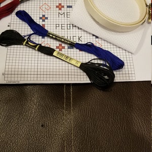 Puede incluir: Un kit de punto de cruz con hilos de bordar de varios colores, una tela blanca y un bastidor de madera. Los colores de los hilos incluyen naranja, burdeos, azul y negro. El texto en el patr&oacute;n dice "ME PEOPLE SUCK".