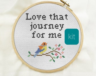 May include: A circular wooden embroidery hoop containing white Aida fabric with a cross-stitch design. The text reads "Love that journey for me" in black thread above a colorful bird perched on a flowering branch with pink and green details.