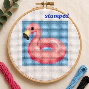 May include: A cross-stitch embroidery hoop featuring a pink flamingo float on a blue background. The word "stamped" is printed above the design. The embroidery is set in a wooden hoop, with pink, blue, and black thread nearby.