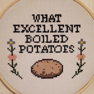 May include: Cross-stitch embroidery in a wooden hoop. The design features the text "WHAT EXCELLENT BOILED POTATOES" in black, with a stitched potato and floral accents. The background is white.