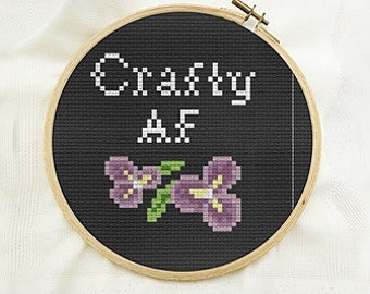 May include: Black embroidery hoop with white text that reads "Crafty AF" and two purple flowers with green leaves.