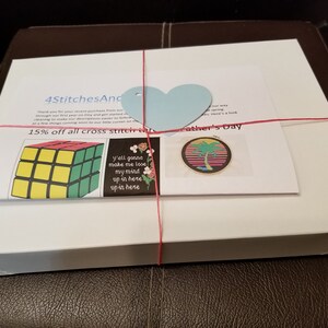 May include: A white box with a red ribbon tied around it. The box is labeled "4StitchesAnd" and has a blue heart-shaped tag. The box contains cross stitch patterns, including a Rubik's Cube, a palm tree, and a floral design. The text "15% off all cross stitch kits" and "Father's Day" is visible on the box.