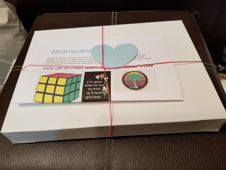 May include: A white box with a red ribbon tied around it. The box is labeled "4StitchesAnd" and has a blue heart-shaped tag. The box contains cross stitch patterns, including a Rubik's Cube, a palm tree, and a floral design. The text "15% off all cross stitch kits" and "Father's Day" is visible on the box.