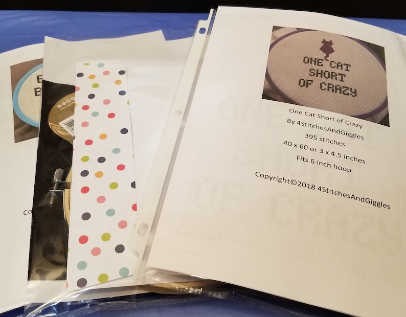 May include: A white sheet of paper with the text "One Cat Short of Crazy" in black lettering. The text is surrounded by a purple and white embroidery hoop. The paper also includes the text "By 4StitchesAndGiggles", "395 stitches", "40 x 60 or 3 x 4.5 inches", "Fits 6 inch hoop", and "Copyright&copy;2018 4StitchesAndGiggles".