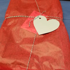 May include: A red gift bag tied with twine and a white heart-shaped tag. The bag is made of tissue paper and has a crinkled texture.