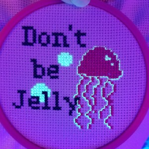 May include: A pink and white embroidery hoop with a cross-stitch design that reads "Don't be Jelly" with a pink jellyfish in the center. The jellyfish is glowing under a black light.