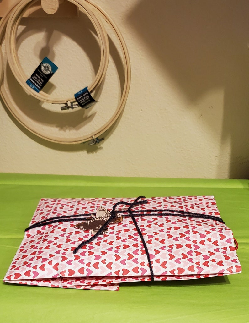 May include: A white gift bag with a red heart pattern is tied with a black ribbon. The bag is sitting on a green surface.